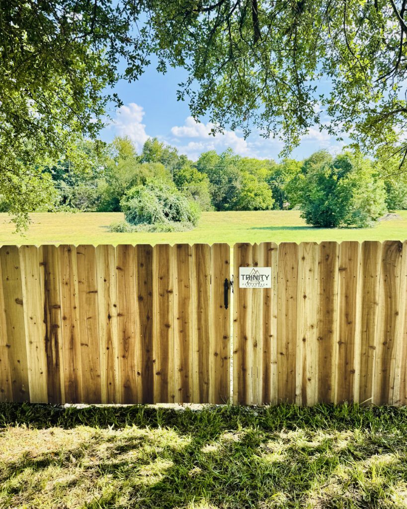 “Trinity Fence & Deck cedar privacy fence under oak tree with company logo plate, overlooking a Texas field”