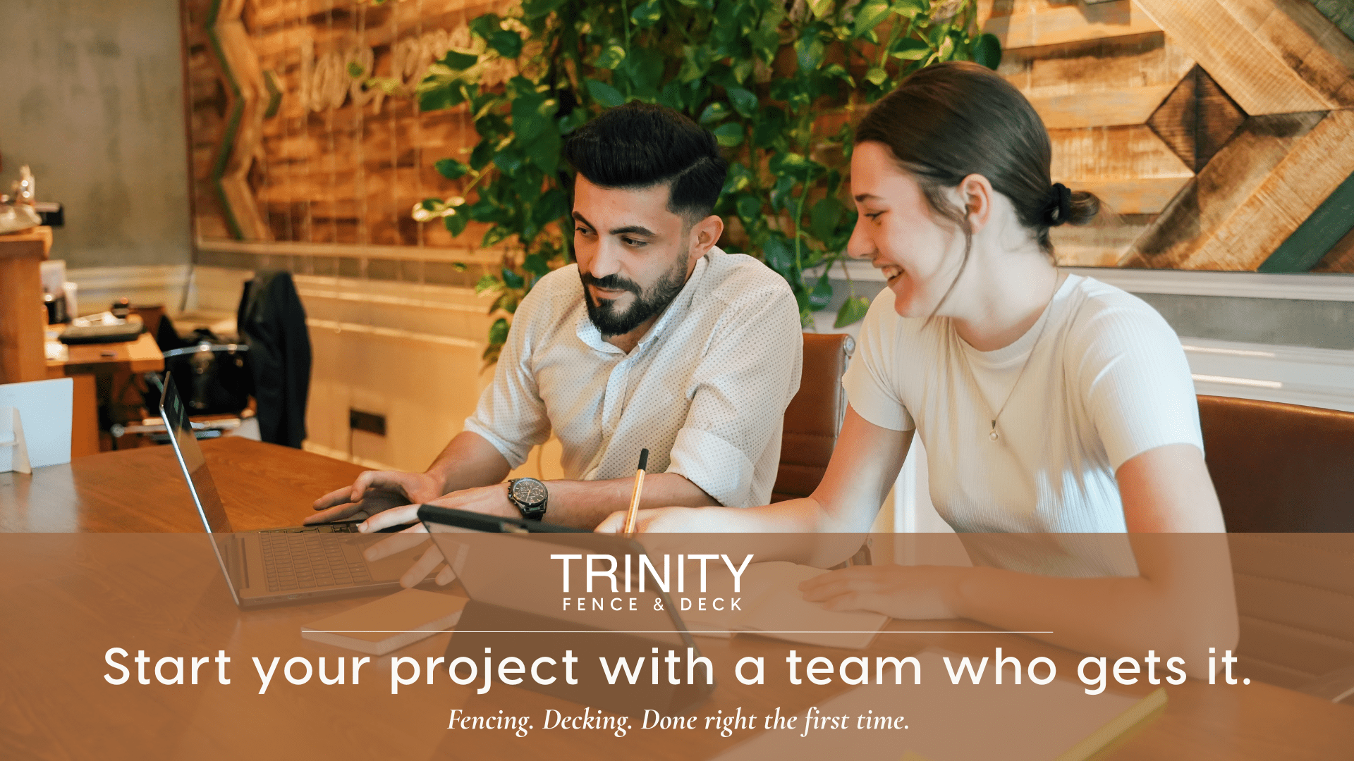 Two people smiling and reviewing a project together at a warm wood table, with the text overlay “Start your project with a team who gets it. Fencing. Decking. Done right the first time.” Trinity Fence & Deck logo centered above.
