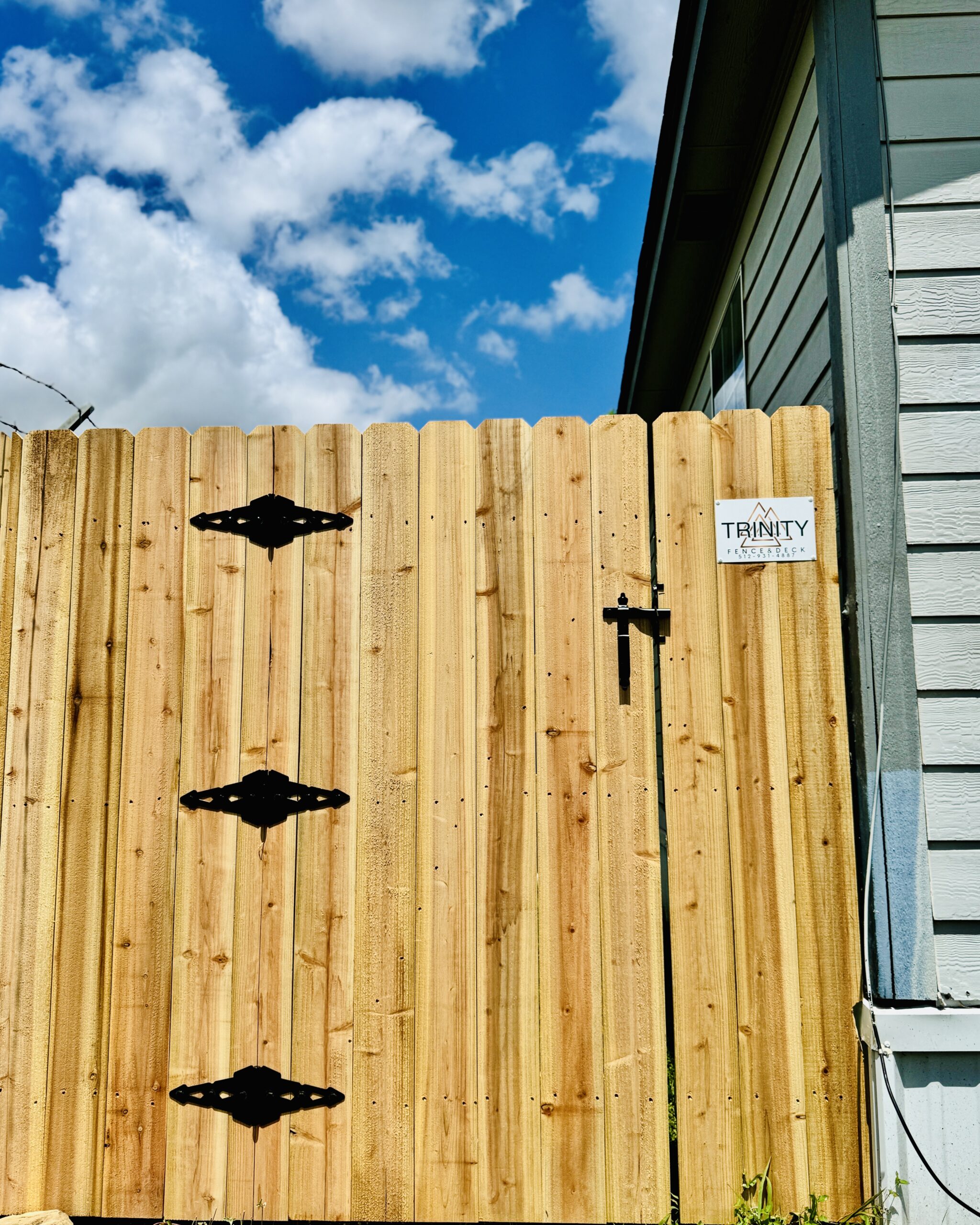 Cedar gate with black hardware and Trinity Fence & Deck branding next to a gray porch