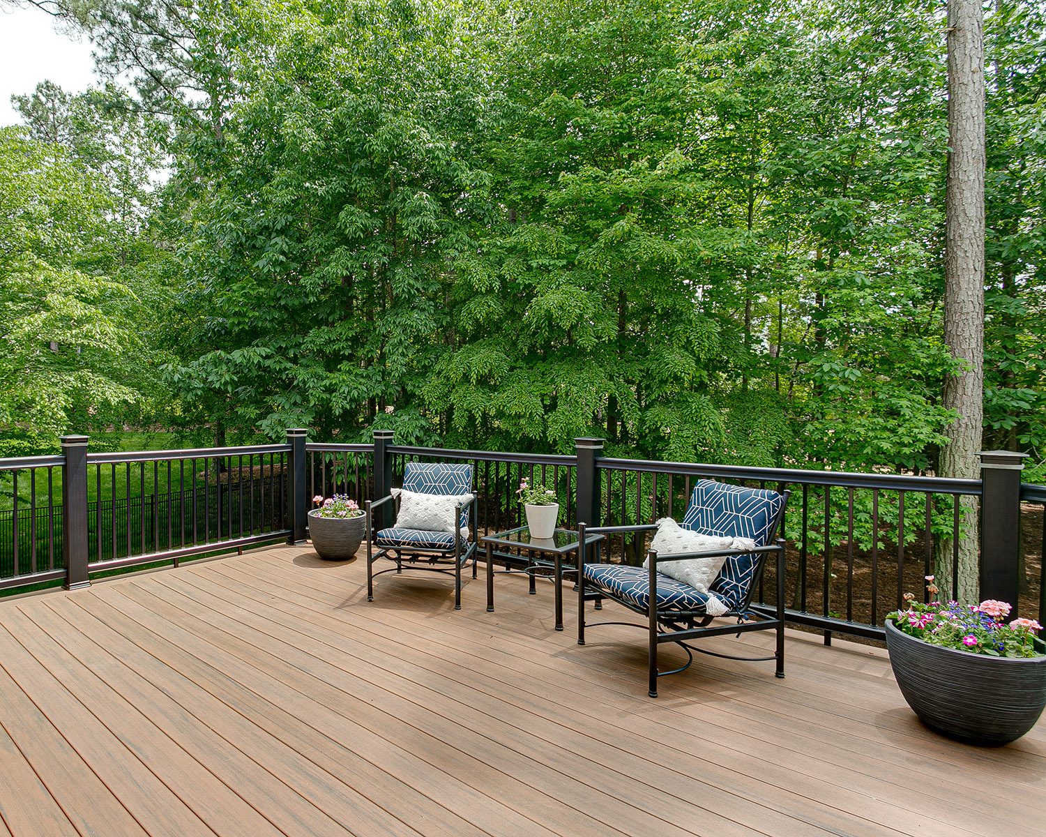 A composite deck with black aluminum railing, two cushioned chairs, a small glass table, and flower planters, set against a backdrop of lush green trees.