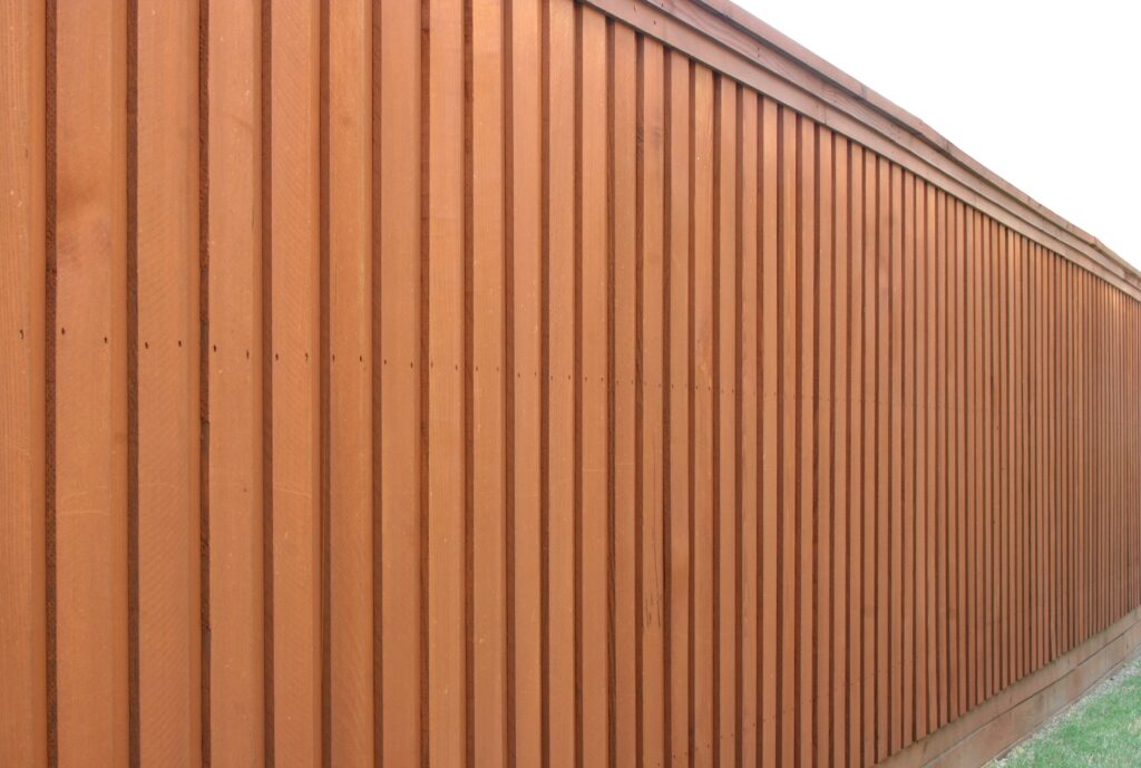 Aged fence shown before stain treatment—ideal for before-and-after comparison Angled view of a tall, stained board-on-board fence with green grass.